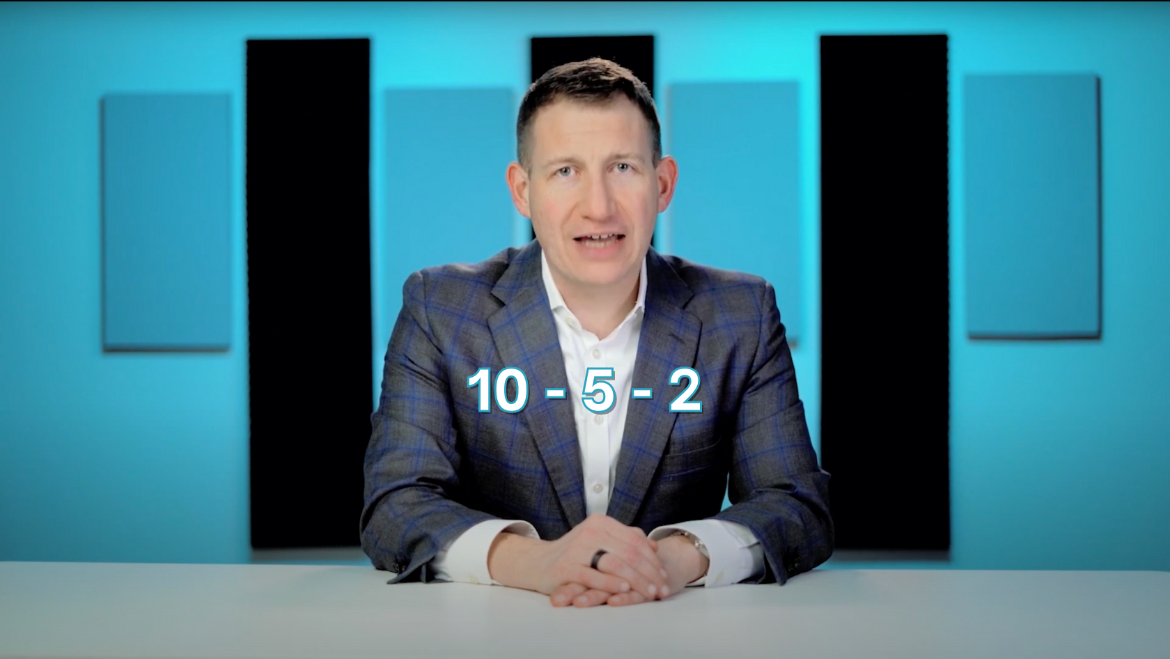 Male presenter in a blue checked blazer at a desk, with the math expression 10 − 5 − 2 on screen.