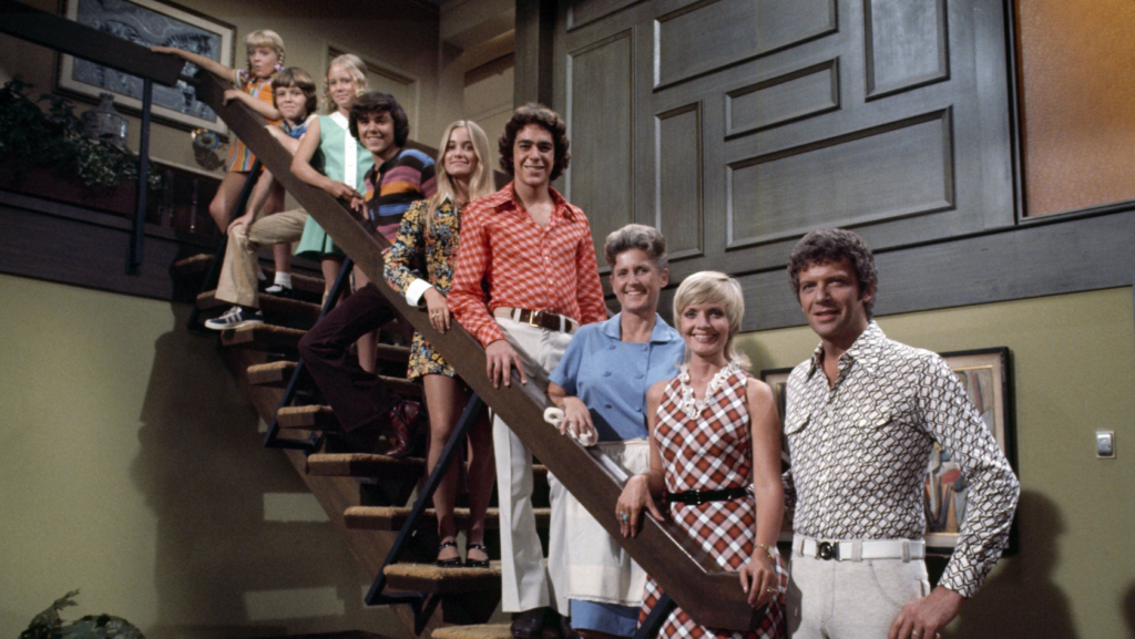 Group of ten people in 1970s outfits posing on a wooden staircase inside a living room with paneled walls and warm colors to create a retro group photo.