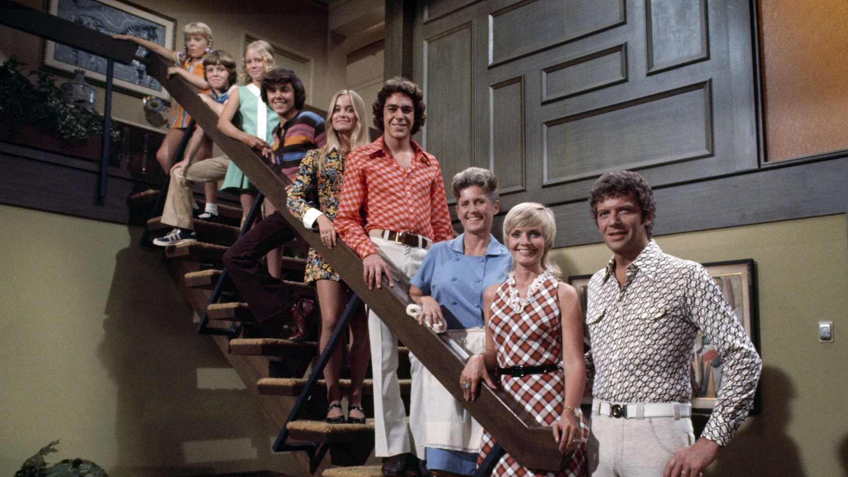 Group of ten people in 1970s outfits posing on a wooden staircase inside a living room with paneled walls and warm colors to create a retro group photo.