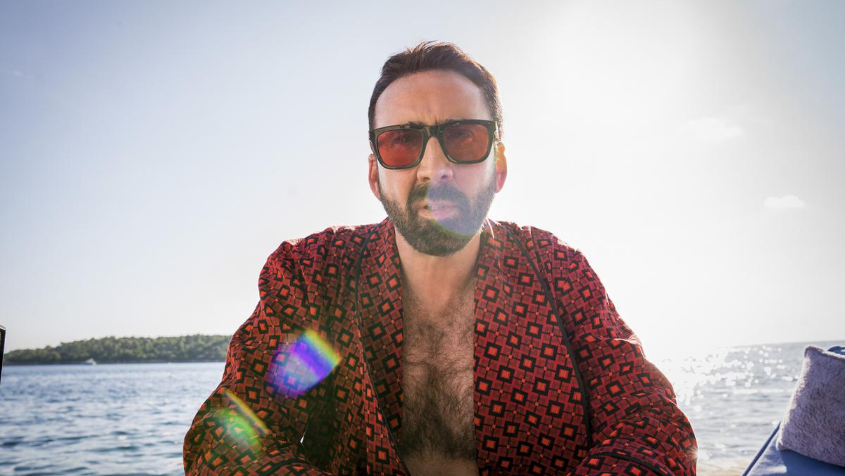 Man with a beard in a red geometric shirt and sunglasses on a boat, sun glare over the water behind him.