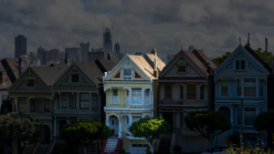 Row of colorful Victorian houses on a steep street with a distant city skyline and cloudy sky behind them.