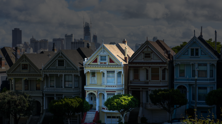 Row of colorful Victorian houses on a steep street with a distant city skyline and cloudy sky behind them.