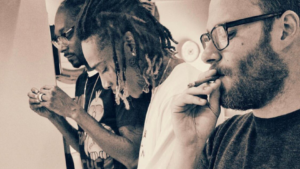 Two men smoking together in a warm, sepia-toned photo; one with dreadlocks and glasses, the other with a beard and glasses in the foreground.