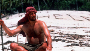 Shirtless man wearing a red headscarf sits on a sandy beach, with large letters formed in the sand behind him and seaweed debris around.