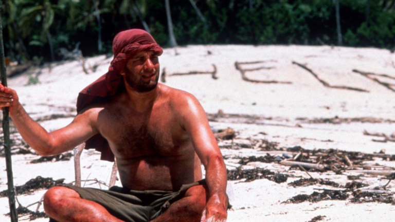Shirtless man wearing a red headscarf sits on a sandy beach, with large letters formed in the sand behind him and seaweed debris around.