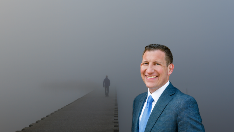 Smiling man in a blue suit takes a shoulder-side portrait against a foggy, misty background with a long, empty walkway to the left
