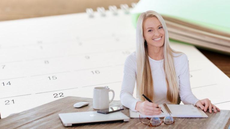 Smiling woman writing in a notebook at a wooden desk, with a large calendar behind her.
