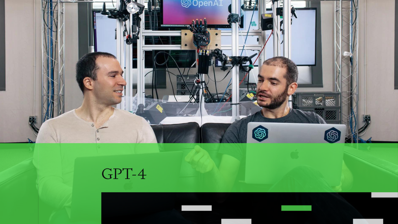 Two men sit on a black couch in a tech lab, each with a laptop, facing and talking to each other amid wired equipment and OpenAI branding behind them.