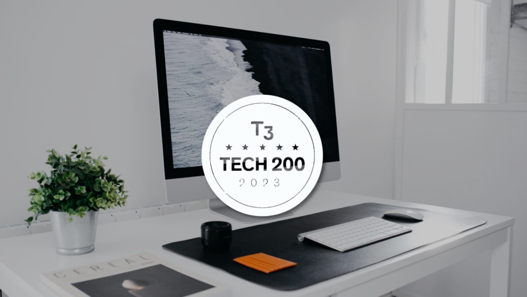 Desk setup with an iMac, keyboard, and plant; a white circular badge reading 'T3 TECH 200 2023' overlays the screen, implying an award.
