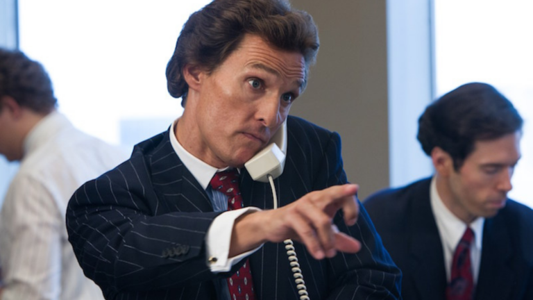Businessman in a pinstripe suit on a landline, pointing as he talks, with colleagues blurred in the background in an office.