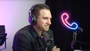 Podcast host wearing over-ear headphones speaks into a microphone in a studio with a neon phone icon in the background (right).