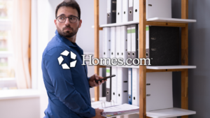 Man in a blue shirt and glasses stands beside a shelf of binders, holding a smartphone; Homes.com watermark across the image.