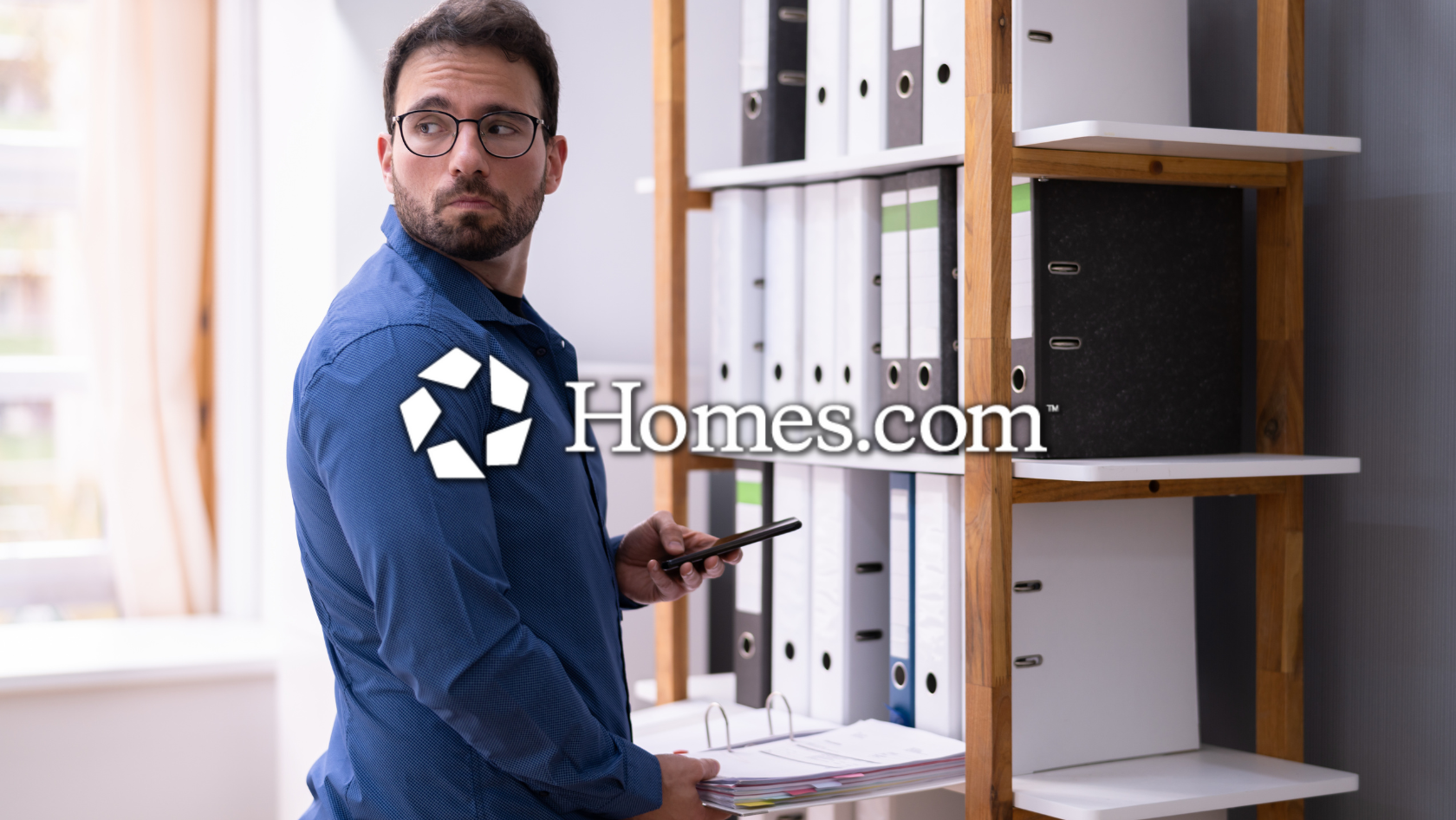 Man in a blue shirt and glasses stands beside a shelf of binders, holding a smartphone; Homes.com watermark across the image.
