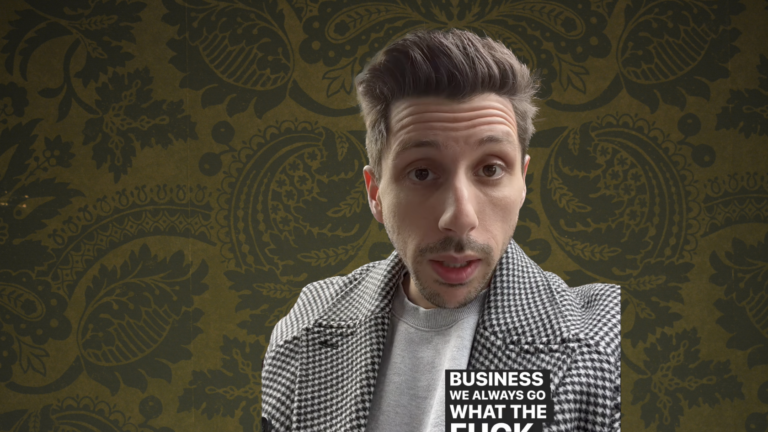Close-up of a man speaking to the camera, wearing a houndstooth coat against a brown patterned wall with a bold text graphic on his torso reading 'BUSINESS WE ALWAYS GO...'.