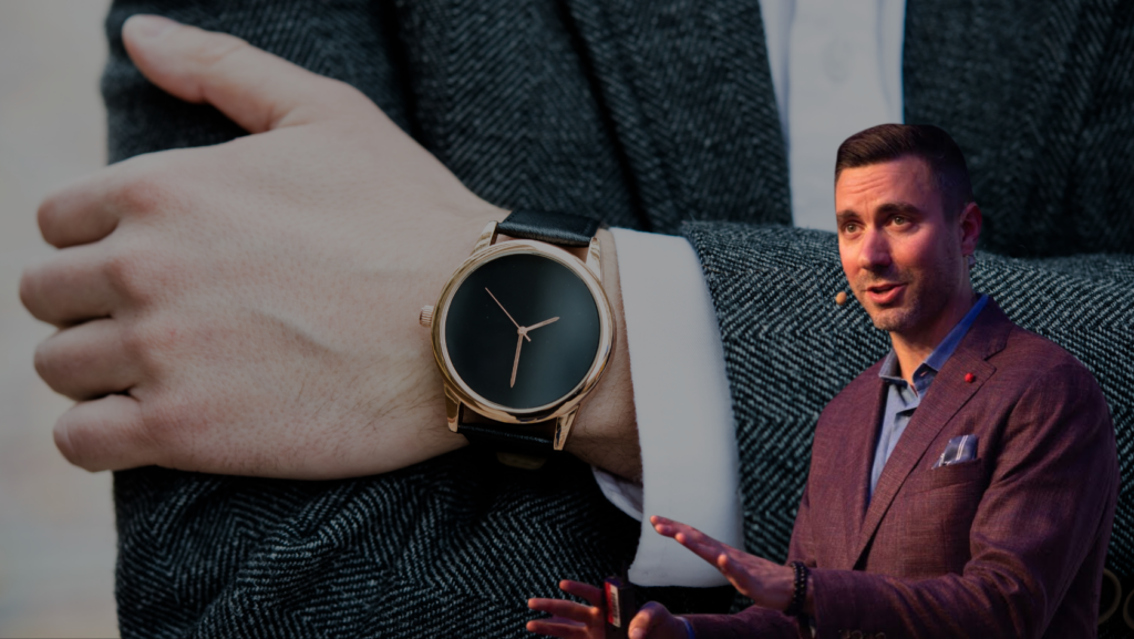 Close-up of a gold wristwatch with a black face on a man's wrist; a suited presenter is visible in the background.