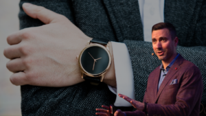 Close-up of a gold wristwatch with a black face on a man's wrist; a suited presenter is visible in the background.
