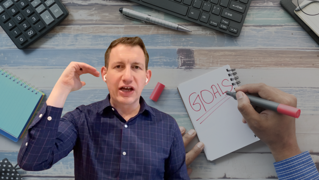 Person speaking to camera with earbuds, desk workspace in background, notebook labeled 'GOALS' and a hand writing with a red marker