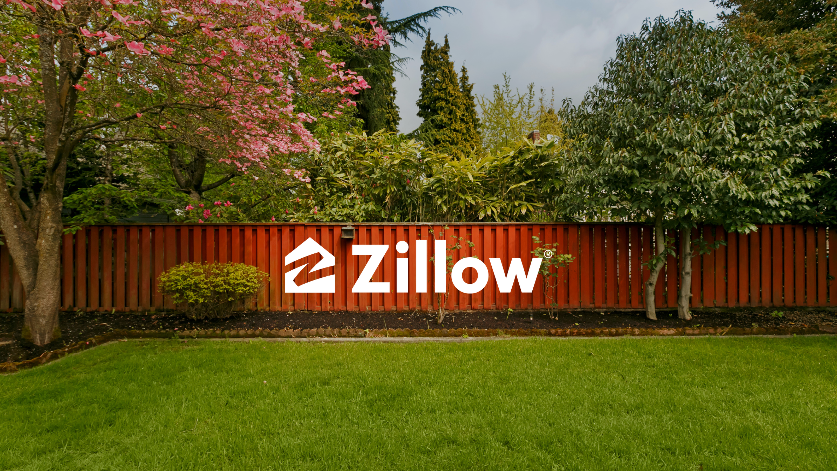 Zillow logo in white on a red wooden fence amid green shrubs and trees in a garden. The lawn is foregrounded.