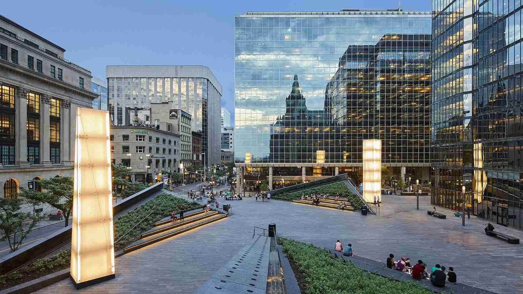 Urban plaza with glass office towers, wide concrete steps, and people walking or sitting nearby.