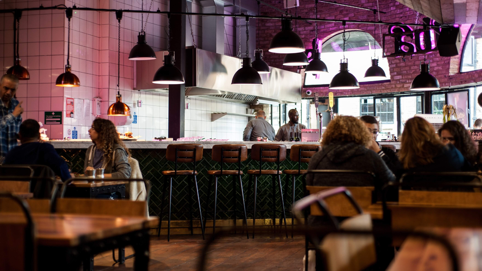 Casual cafe interior with a long bar, row of brown stools, and warm pendant lights; diners sit at tables in the foreground while staff work behind the counter.