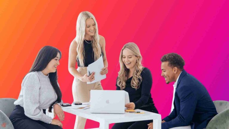 Four professionals collaborate around a white table: a laptop open, papers in hand, against a bright orange-pink gradient background.
