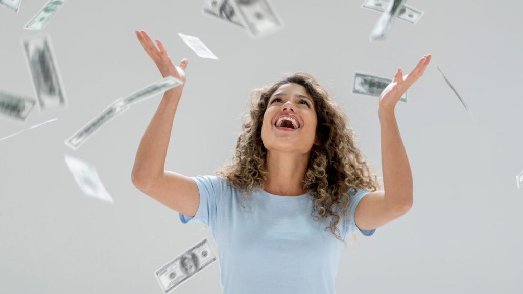 Joyful young woman in a light blue shirt raises her arms as dollar bills float around her.
