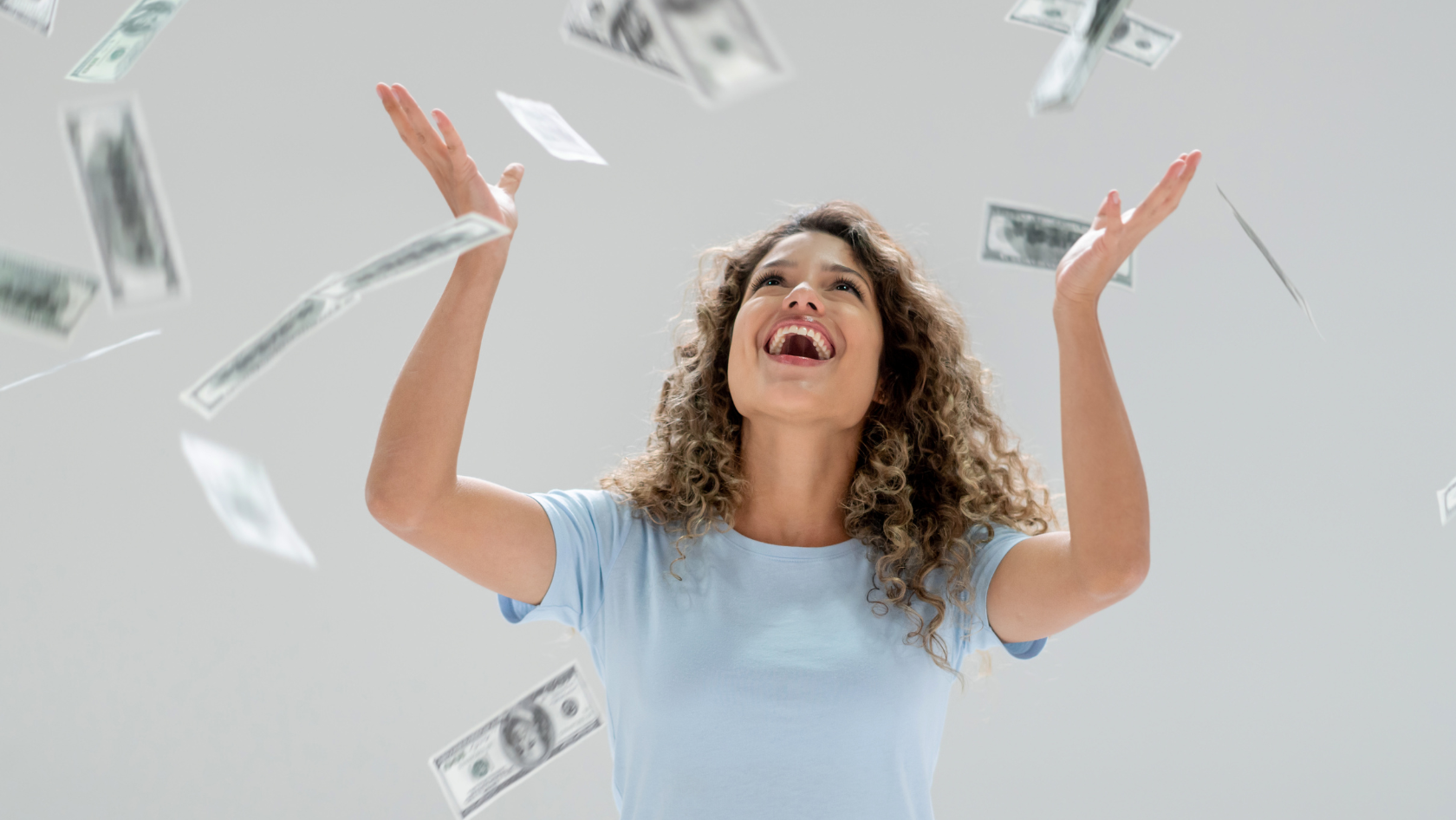 Joyful young woman in a light blue shirt raises her arms as dollar bills float around her.