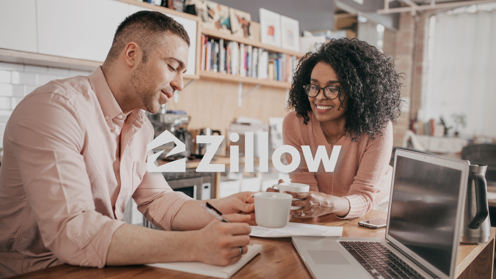 Two colleagues discuss documents over coffee at a wooden table, with a laptop nearby and a Zillow watermark overlaying the image.
