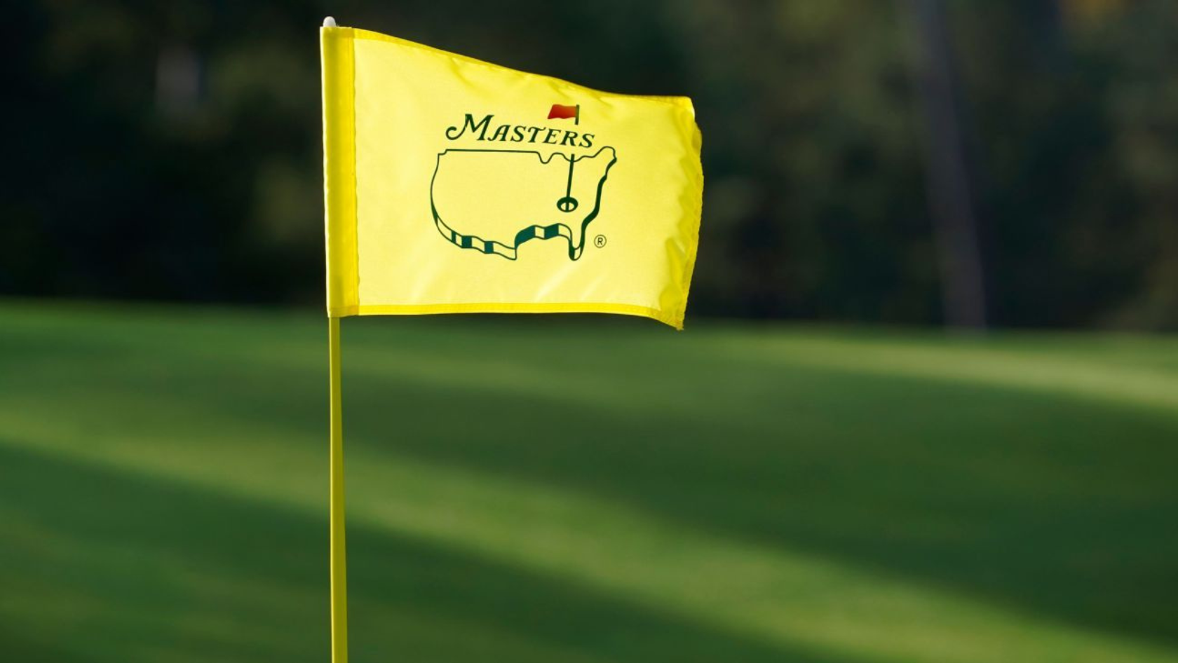 Yellow Masters flag on a golf green flagstick, with the Masters logo and name visible against a blurred grassy background.