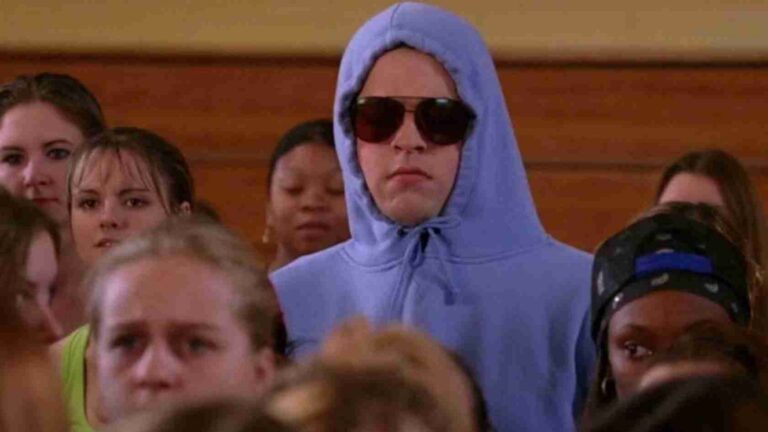 A person wearing a light blue hoodie and dark sunglasses stands among a crowd indoors.