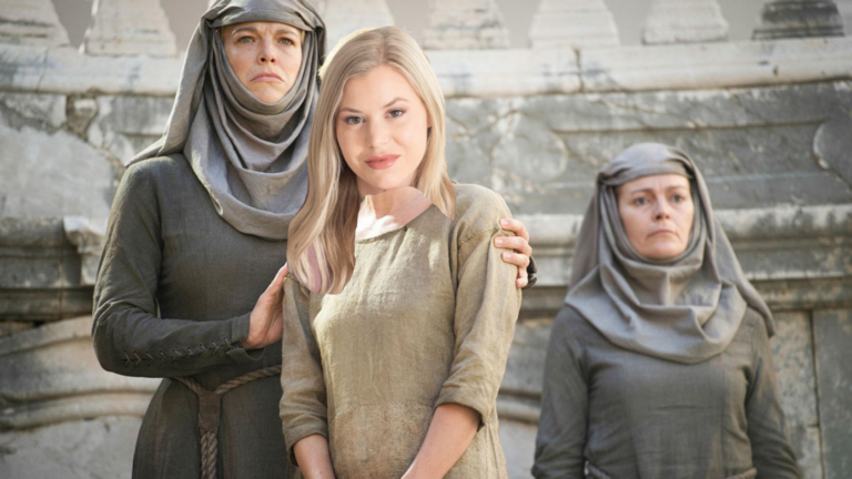 Three women in medieval-style robes pose in front of a stone ruin; a blonde woman in beige stands center with two gray-robed women beside her, one touching her shoulder.