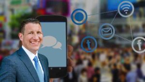Smiling businessman in a suit holds a smartphone showing chat bubbles, with floating social media icons in the background.