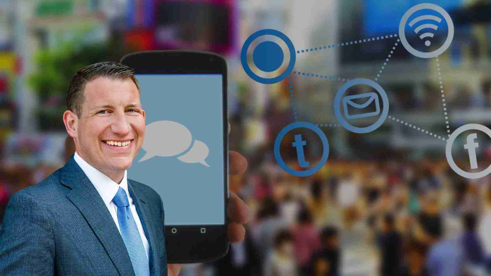 Smiling businessman in a suit holds a smartphone showing chat bubbles, with floating social media icons in the background.