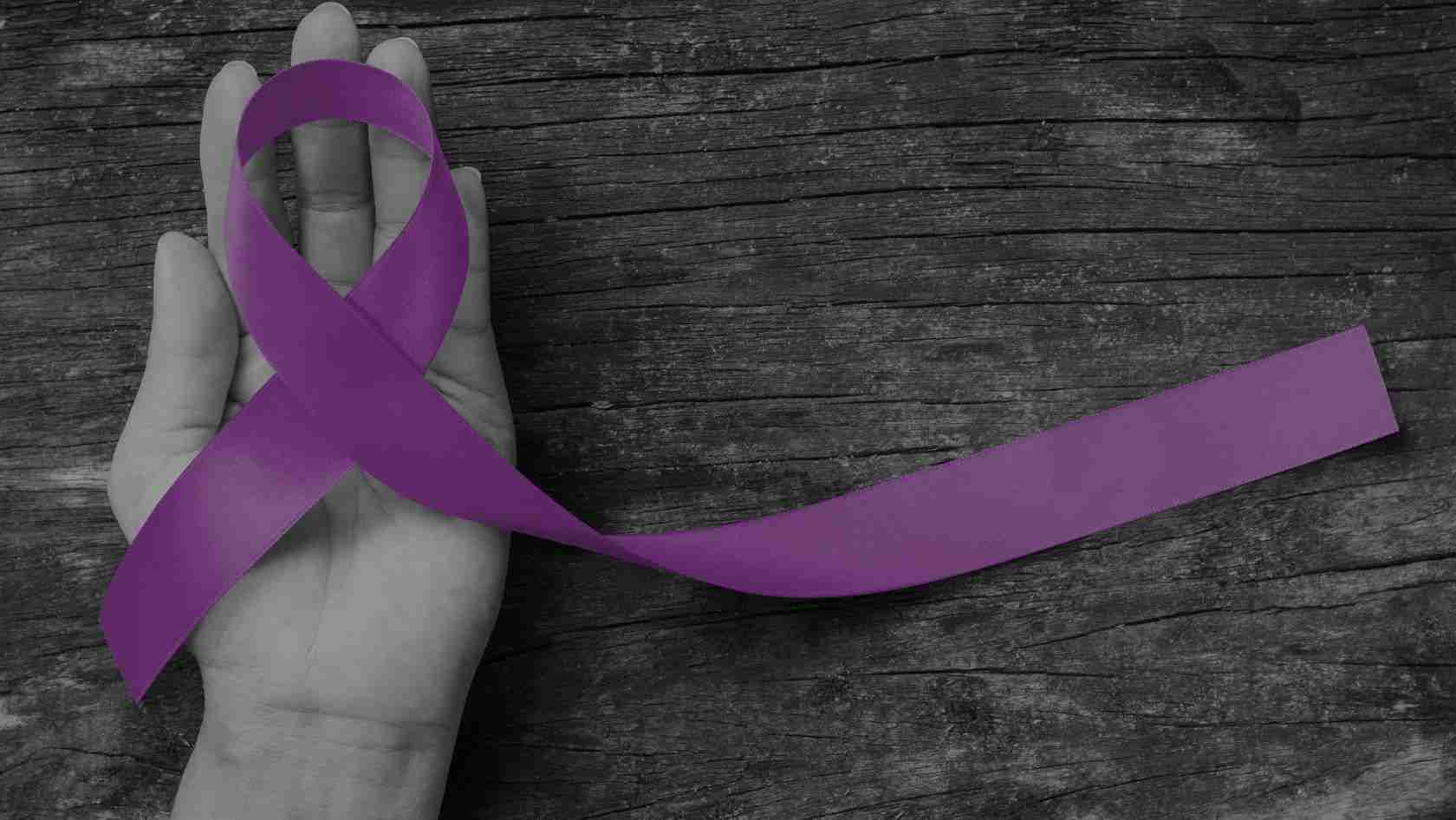 Purple awareness ribbon draped over an open hand on a dark wooden background, symbolizing support for a cause.