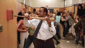 A man in a dress shirt and tie swings a wooden bat at a wall in a crowded school hallway.