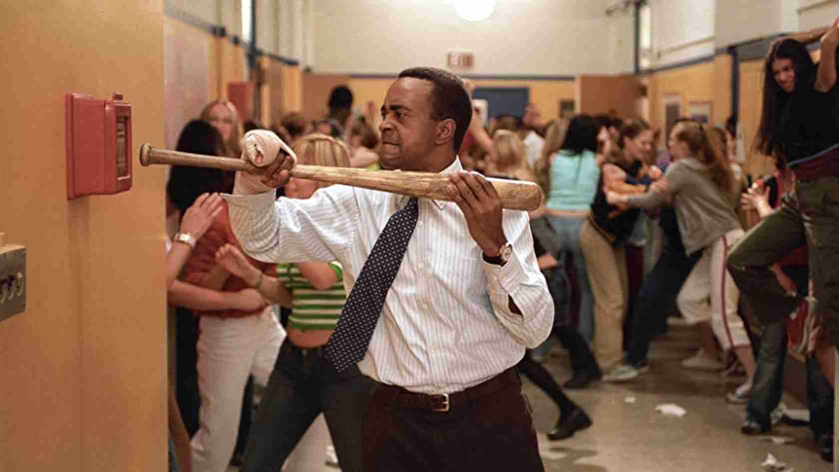 A man in a dress shirt and tie swings a wooden bat at a wall in a crowded school hallway.