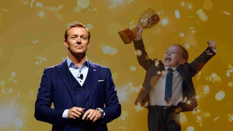 Man in a navy suit stands with hands clasped as a young boy in a suit raises a trophy amid gold confetti on stage.