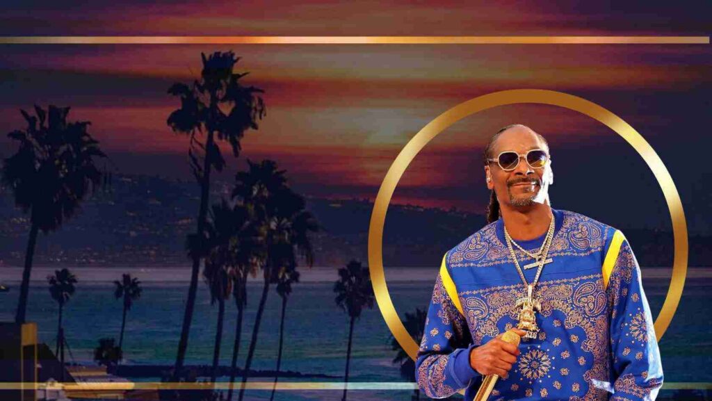 Male performer in a blue patterned shirt with gold chains stands before a sunset beach backdrop with palm trees and a gold circular frame.