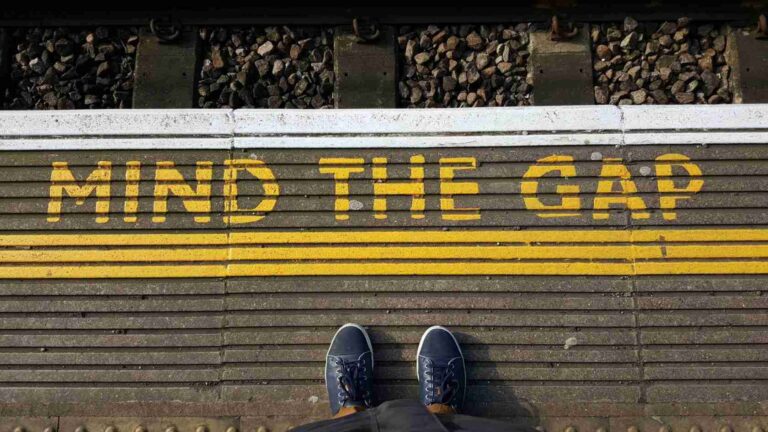 Top-down view of a person standing on a platform with bright yellow 'MIND THE GAP' warning text along the edge of the tracks.
