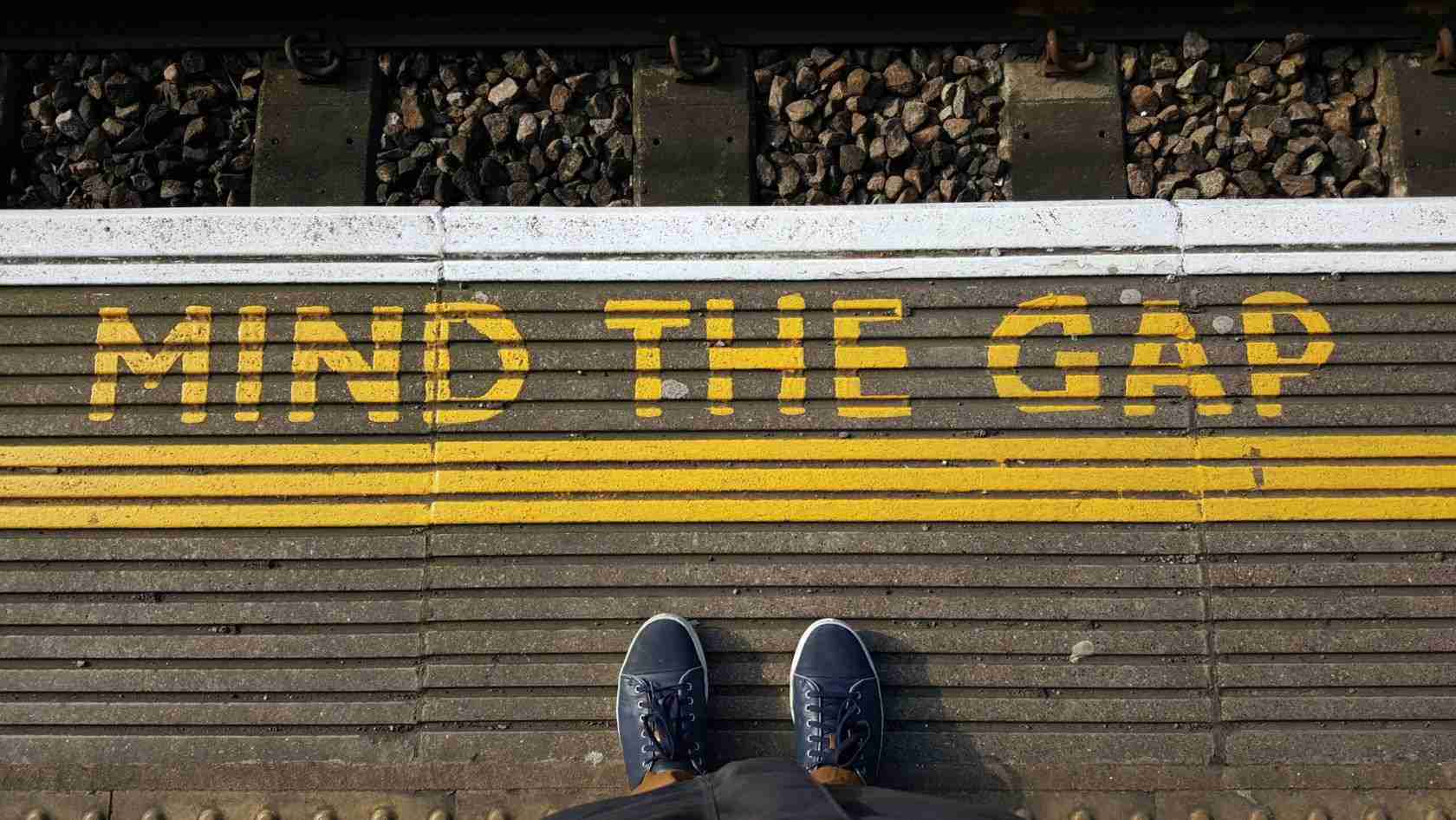 Top-down view of a person standing on a platform with bright yellow 'MIND THE GAP' warning text along the edge of the tracks.