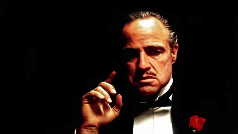 Serd, older man in a black tuxedo with a white shirt and red rose boutonniere, raising his index finger against a dark background.