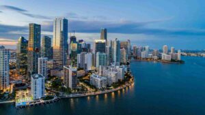 Aerial view of a downtown city skyline along a curved shoreline at dusk, with tall glass buildings and calm blue water reflecting lights.