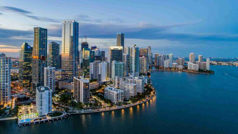 Aerial view of a downtown city skyline along a curved shoreline at dusk, with tall glass buildings and calm blue water reflecting lights.