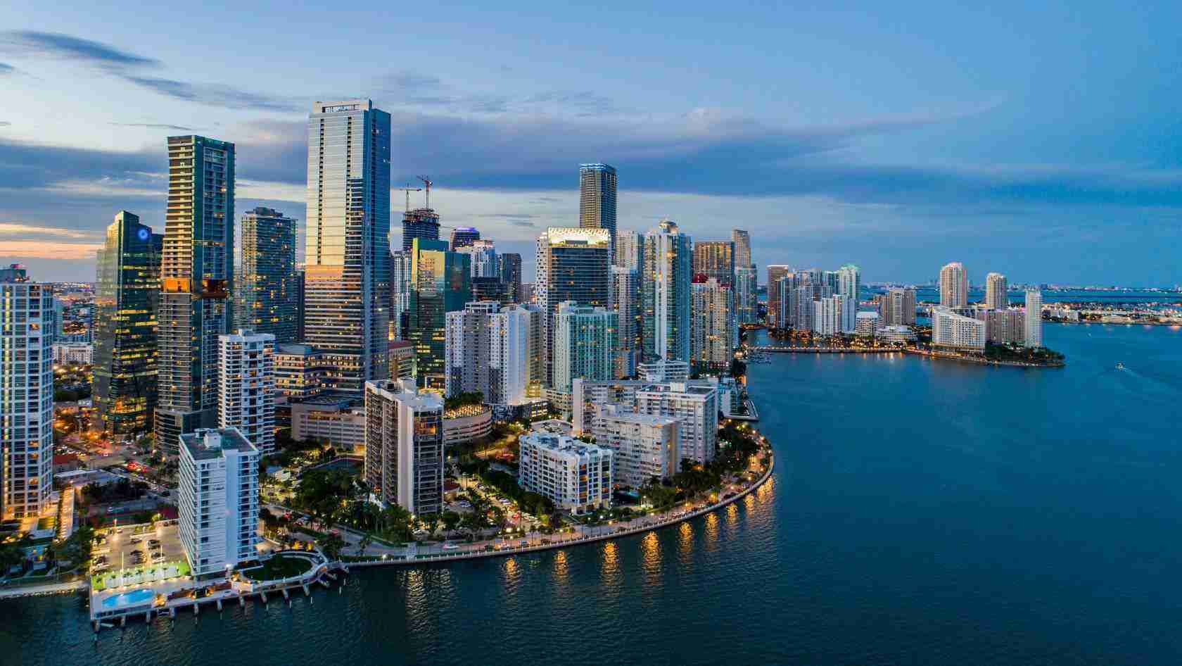 Aerial view of a downtown city skyline along a curved shoreline at dusk, with tall glass buildings and calm blue water reflecting lights.