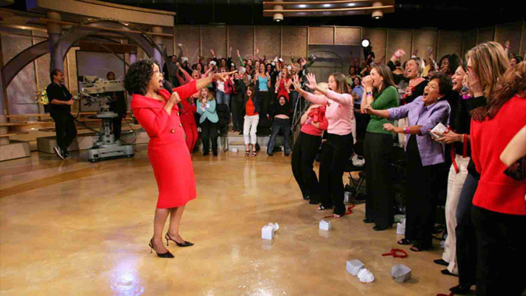 Female host in a red dress sings to a cheering studio audience reaching out toward her.