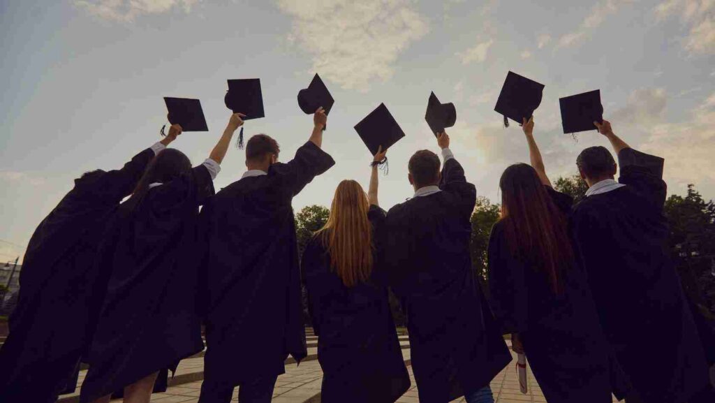 Graduates in black gowns throwing mortarboard caps in the air during an outdoor celebration at sunset/afternoon.
