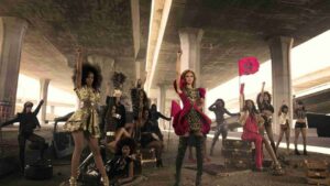 Group of fashion models posing with raised fists under a highway overpass, wearing bold metallic and patterned outfits with props nearby.”