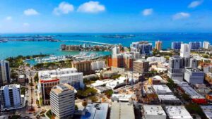 Aerial view of a sunny coastal city with high-rise buildings, a marina full of boats, and a long bridge over turquoise water.