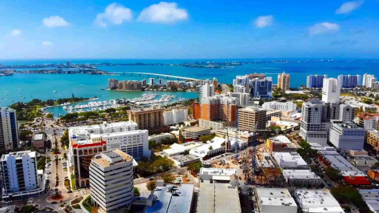 Aerial view of a sunny coastal city with high-rise buildings, a marina full of boats, and a long bridge over turquoise water.