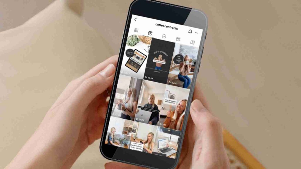 Person's hands hold a smartphone showing a grid view of video posts (reels) on a social media app against a beige background.